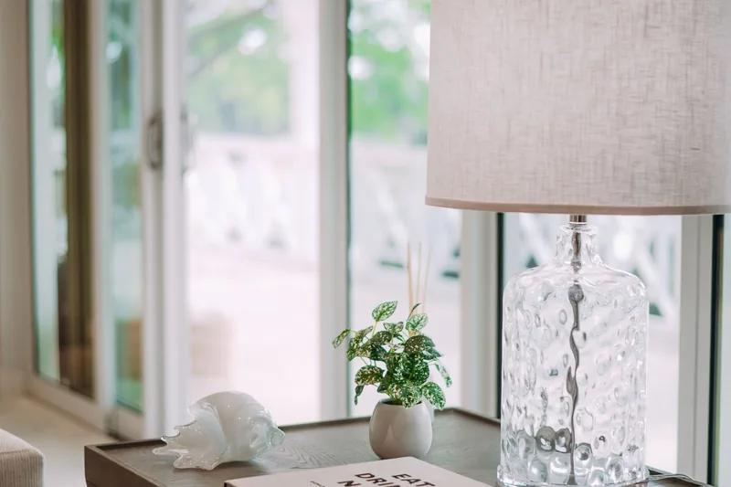 A glass lamp base and a shell on a side table beside an open window