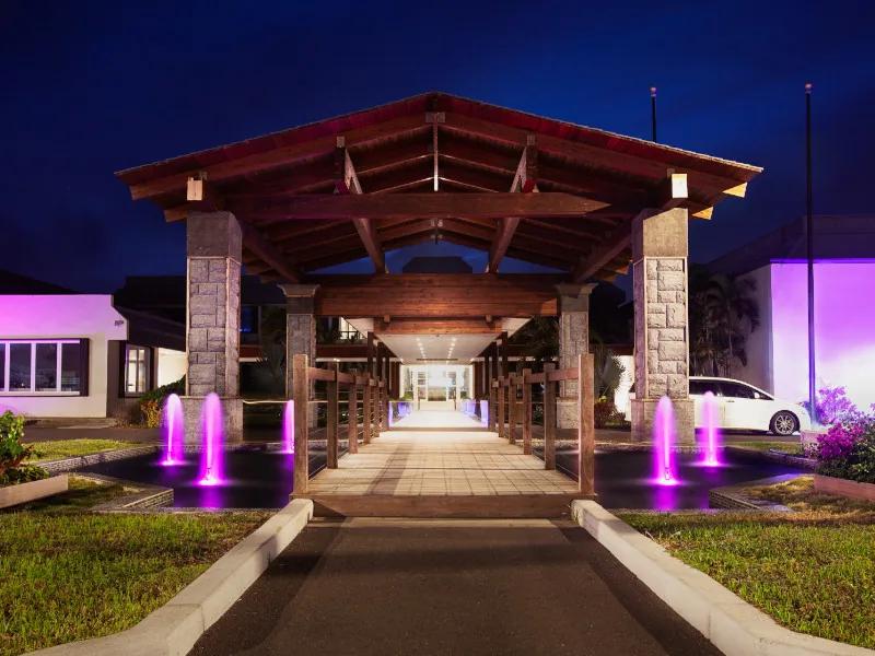 A timber porte-cochere lit at night, magenta-lit fountains along the drive