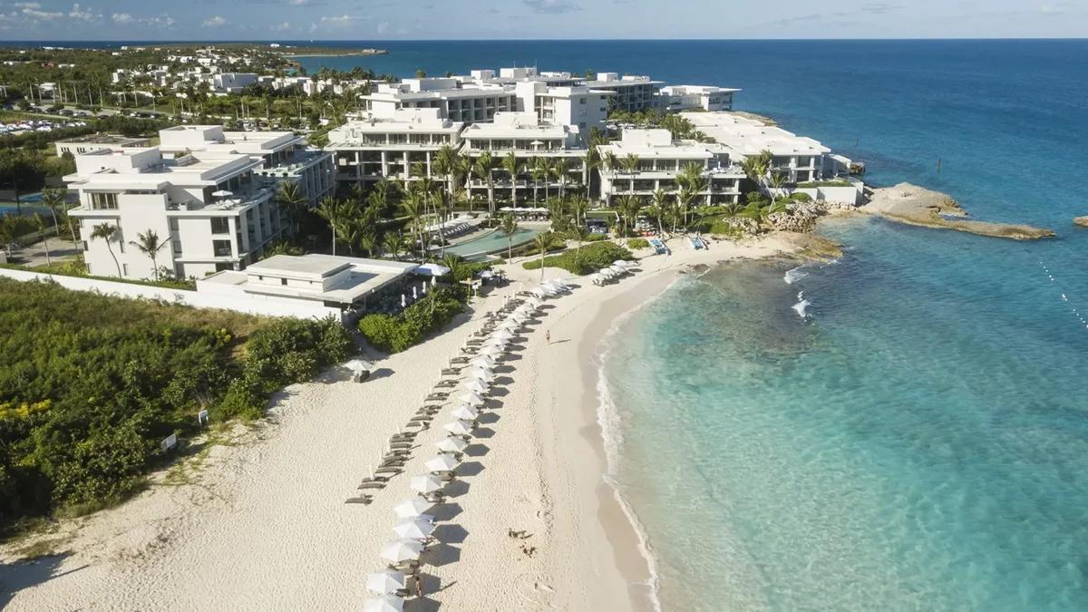 A white resort on a low headland above a beach lined with parasols and loungers
