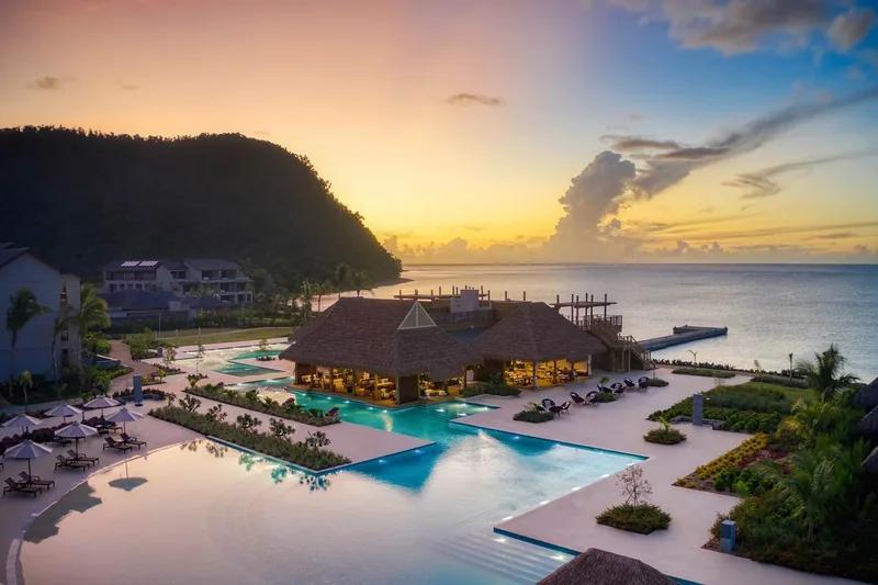A dusk aerial view of a resort pool complex on Dominica's Douglas Bay, a thatched pavilion and a headland silhouetted against a sunset sky
