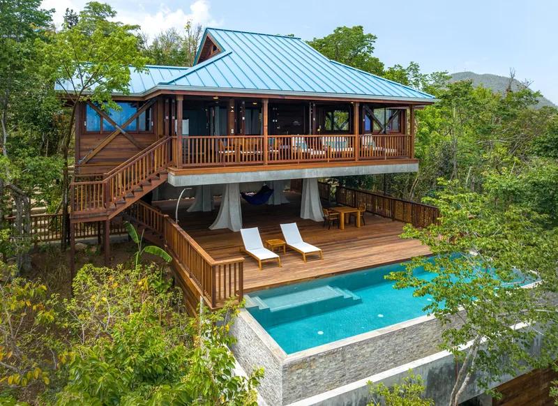 A timber villa with a blue metal roof, its deck and plunge pool above the forest