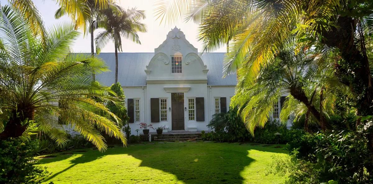 A white house with a curved Dutch gable and green lawn, framed by palms