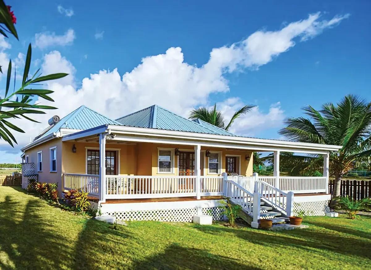 A yellow timber house with a blue metal roof and a wide covered verandah, standing on a lawn behind palms