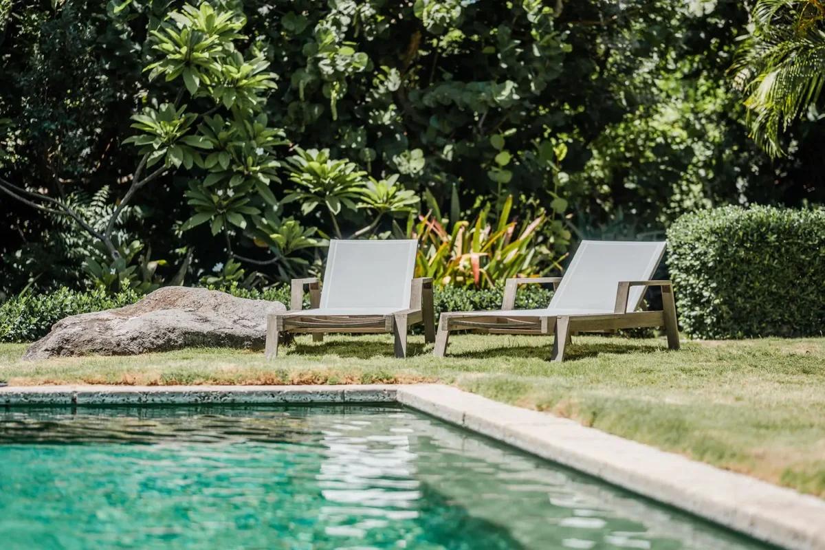 A pool with two loungers on a lawn, dense greenery and boulders behind