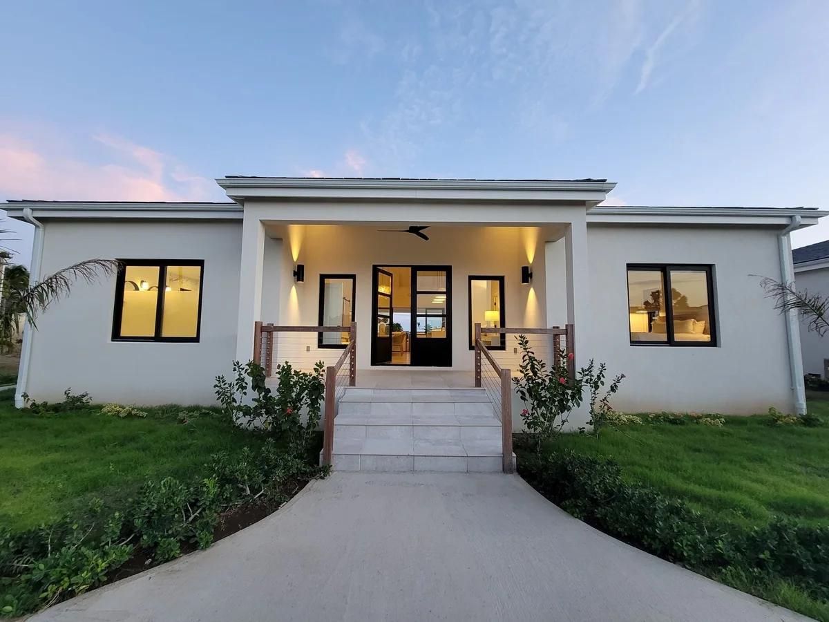 A white single-storey villa at dusk, its lit entrance at the head of a paved path across the lawn