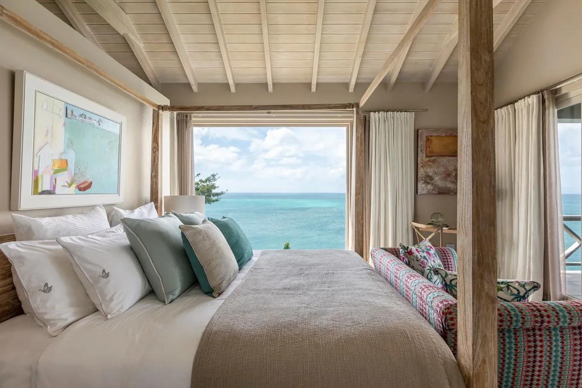 A bedroom under a white timber ceiling with a window framing the sea