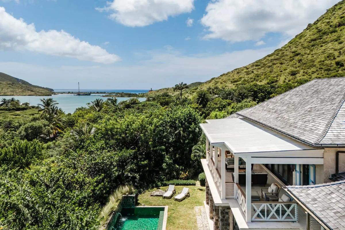 A shingle-roofed villa on a green hillside above a bay, its pool and loungers on the terrace below
