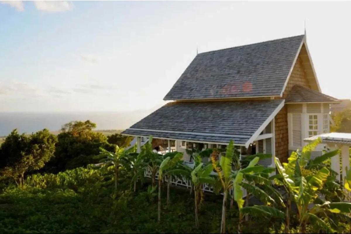 A steep-roofed timber cottage on a green hillside in St Kitts and Nevis, banana plants in the foreground and the sea beyond