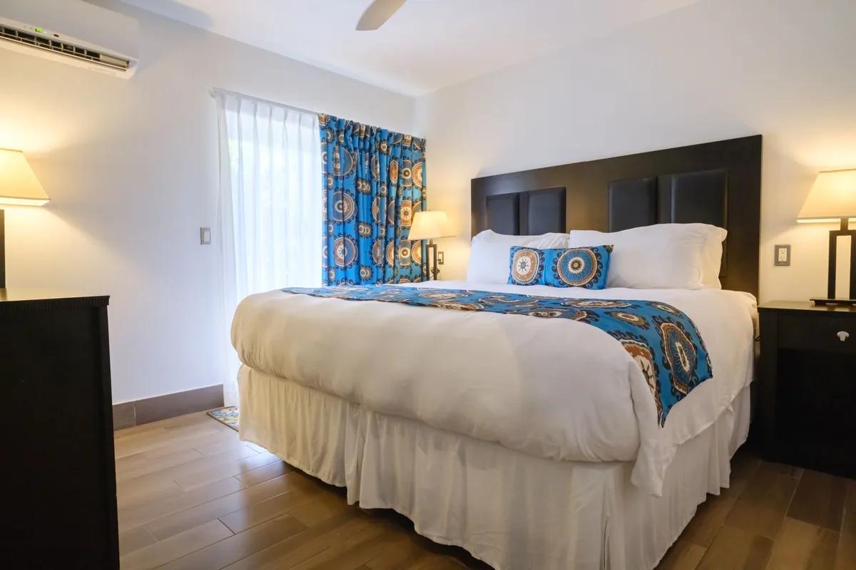 A furnished bedroom in a completed condominium at the Royal St Kitts, with a king bed, dark headboard, ceiling fan and a curtained window onto the grounds