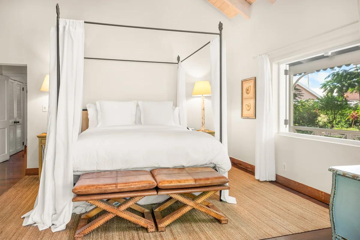 A four-poster bed draped in white linen on a timber floor, a garden window alongside