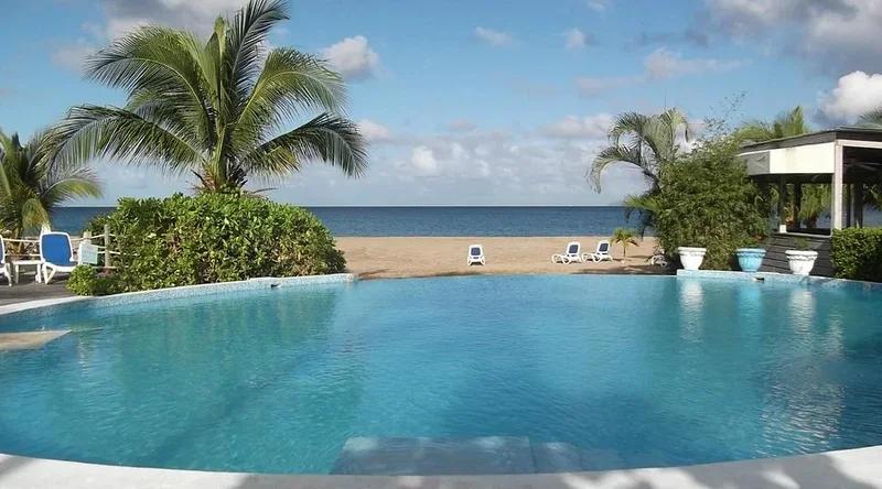 A wide pool beside a beach, palms and low buildings along the shore