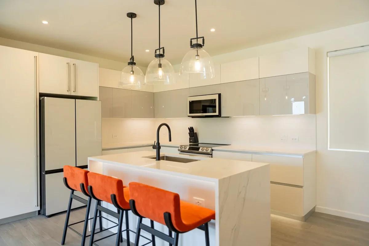 A white kitchen island with orange bar stools and three glass pendant lights