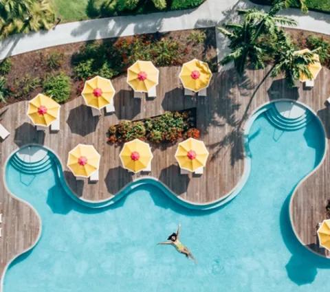 A curving pool with yellow parasols seen from above, a swimmer floating in the clear water