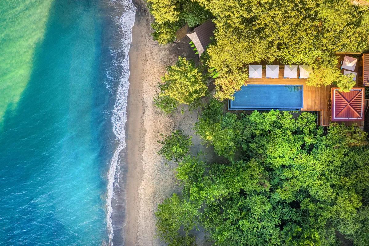 Overhead aerial of a waterfront villa pool deck set in rainforest above Secret Bay's shoreline, Dominica