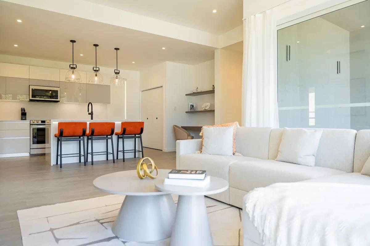 A white living room with a sectional sofa, the kitchen and orange bar stools beyond
