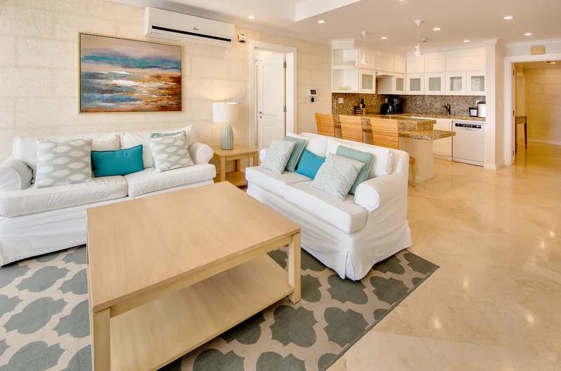 A white living room with turquoise cushions on a patterned rug, an open kitchen behind
