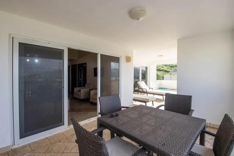 A covered terrace with a dark wicker dining table and chairs beside sliding glass doors