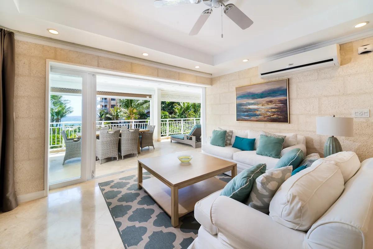 A cream-toned living room under a ceiling fan, opening through wide doors to a covered terrace and the sea