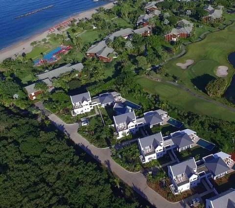 White gabled villas among trees between a golf course and the beach, seen from the air