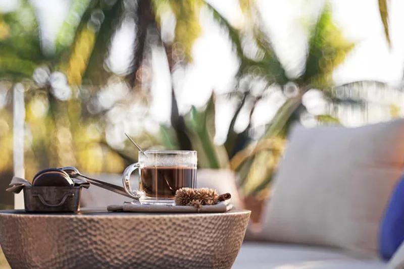 A glass cup of coffee on a woven tray, palms out of focus behind