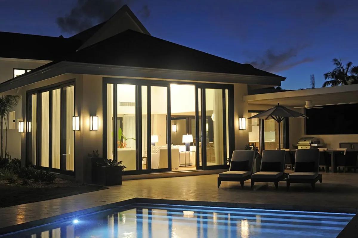 A single-storey villa lit from within at dusk beside a pool and loungers