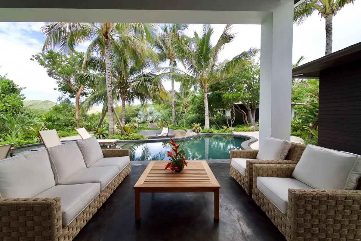 A covered lounge of cushioned sofas looking out to a pool framed by palms