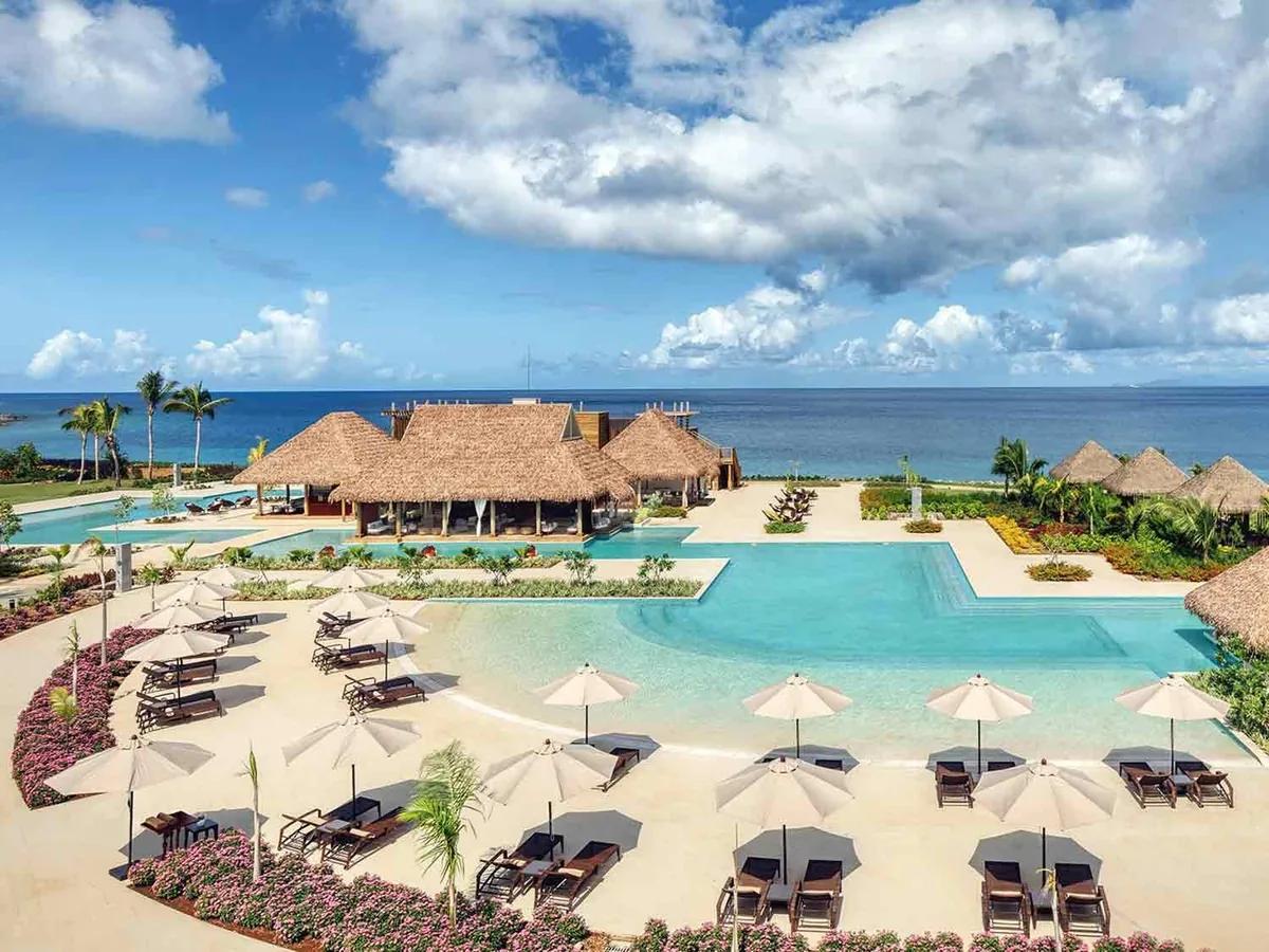 An aerial view of a resort's beachfront pool complex in Dominica, a thatched pavilion, loungers and umbrellas on the pool terrace, and the sea beyond