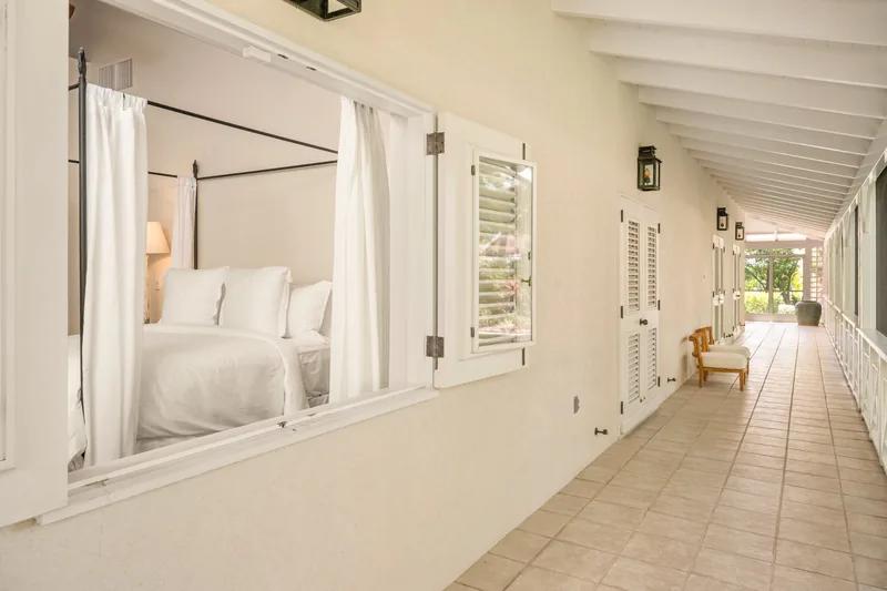 A white four-poster bed seen from a long tiled corridor with louvred doors