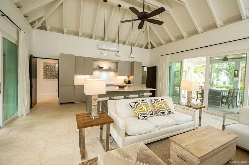 A living room under a white vaulted ceiling with a dark ceiling fan, white sofa and open kitchen behind