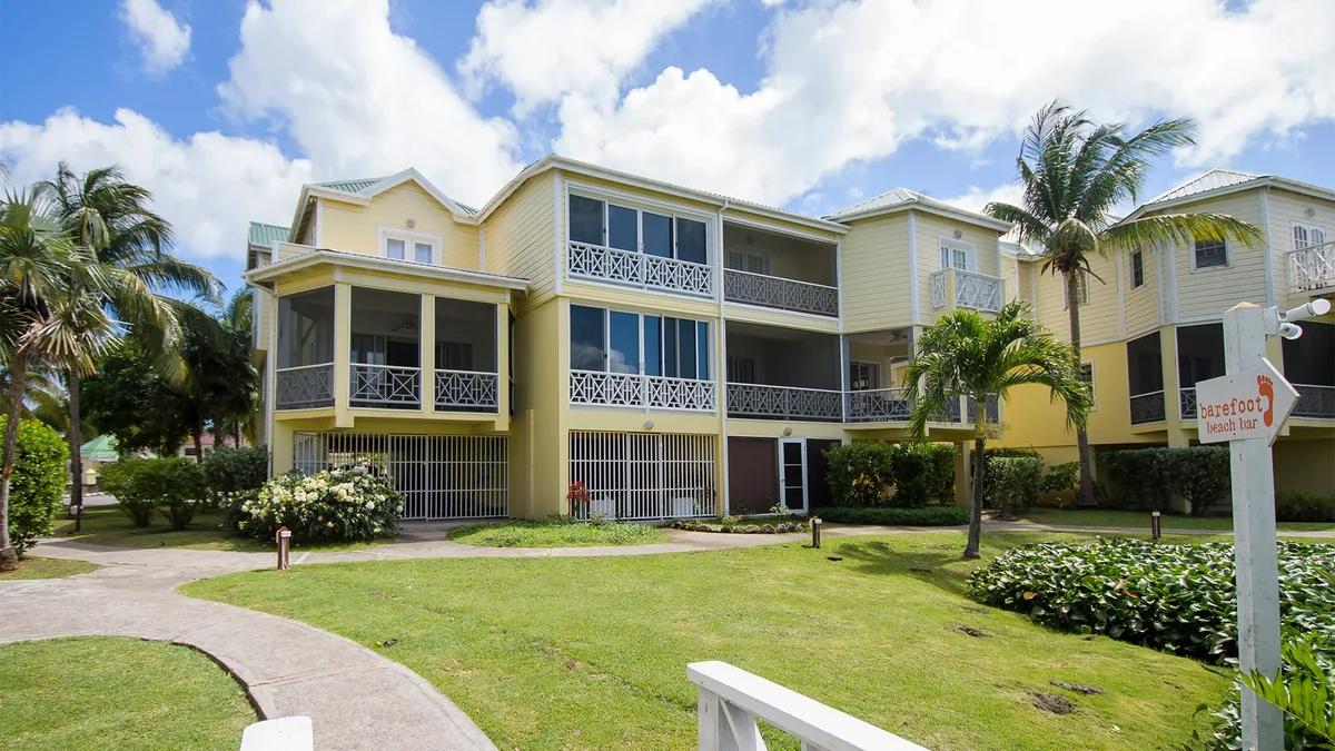 A yellow apartment building with screened balconies behind a lawn and palms