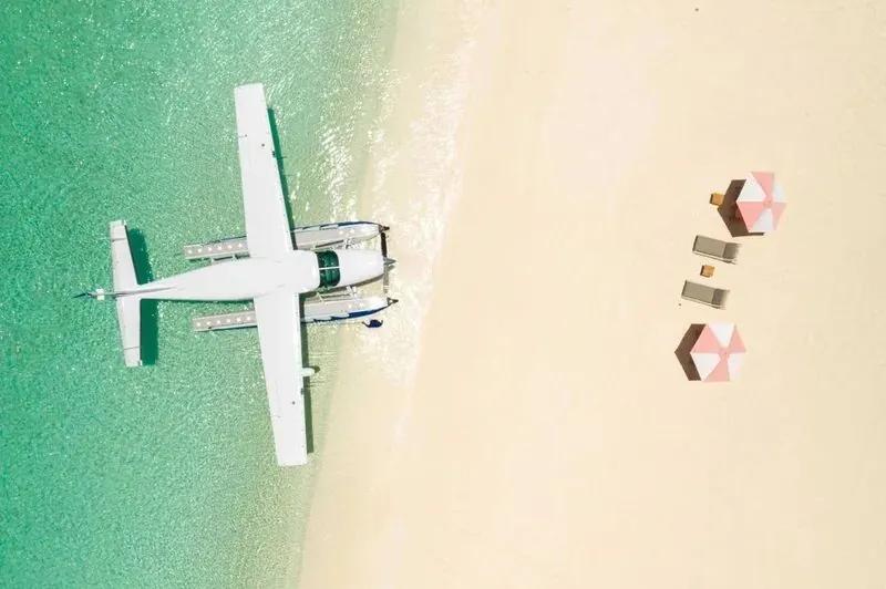 A light seaplane parked on pale Barbuda sand at the water's edge, seen from directly above, beach parasols alongside