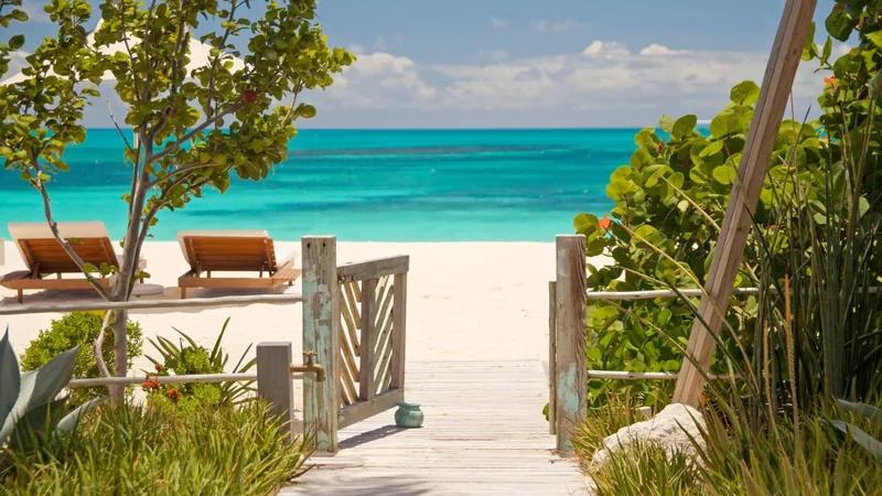 A timber gate opening from planting onto a white beach, two loungers set under a tree