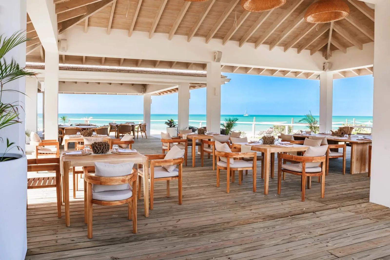 A covered restaurant terrace of timber tables and chairs looking out to turquoise water
