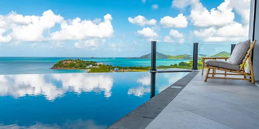 An infinity pool reflecting the sky, a single chair at its edge and an island on the horizon