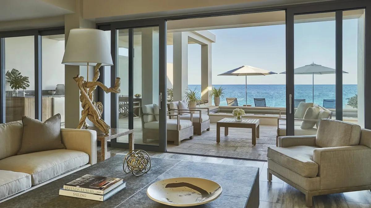 A pale living room opening through sliding glass to a terrace with parasols and the sea beyond